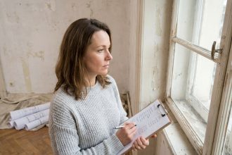 Femme tenant un devis budgétaire devant une vieille fenêtre en bois à rénover dans un salon